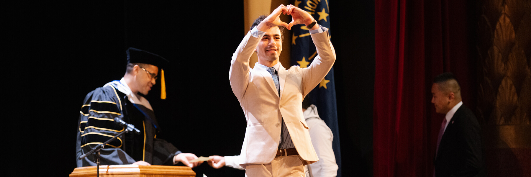 A student on stage making a heart sign with their hands at a commencement ceremony.