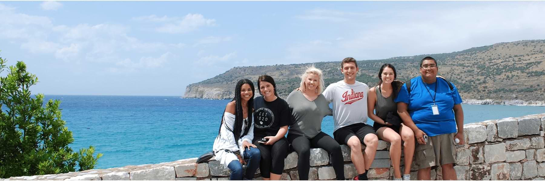 A group of students studying abroad in Greece pose for the camera by a stone wall near the ocean.